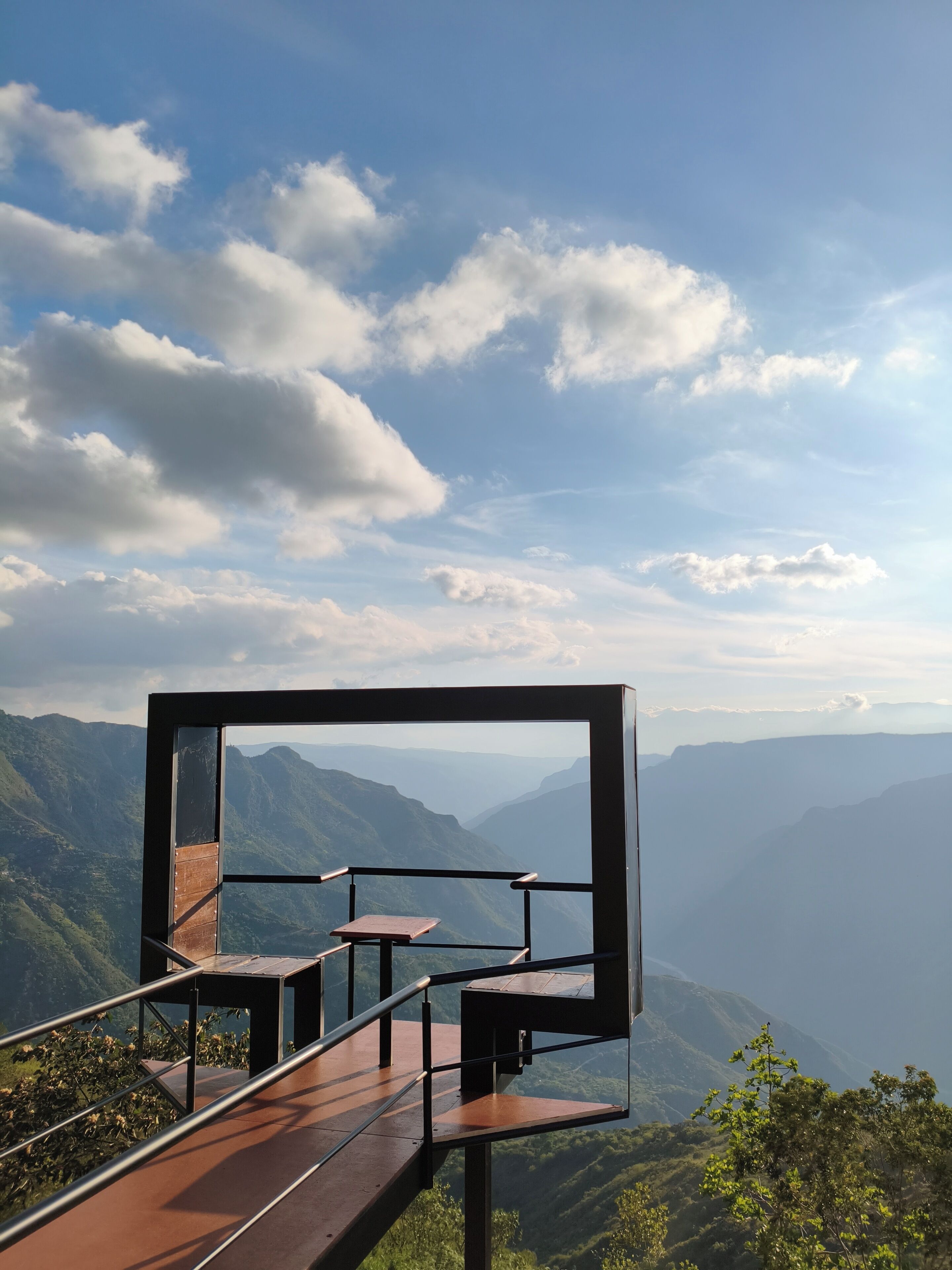 Vista panorámica desde un lugar elevado que domina un valle con montañas, cielo despejado y una moderna plataforma de observación con bancos.
