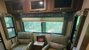Een gezellig interieur van een camper met twee leren fauteuils, een kleine tafel en een kachel, en ramen met uitzicht op groen.