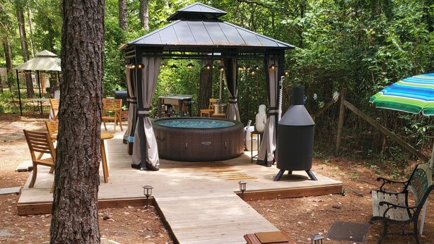 Een houten terras leidt naar een pergola met een jacuzzi, omringd door tuinmeubels en een grote barbecue.