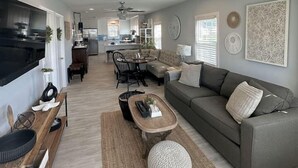 Un salon moderne avec un canapé, une table basse et un téléviseur. On y trouve également un coin repas avec des chaises et une table. La pièce est décorée dans des tons neutres et on aperçoit une cuisine au fond.