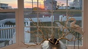 Une pièce avec une grande fenêtre, un vase en métal orné de branches et, à l’extérieur, une clôture blanche faite de piquets.