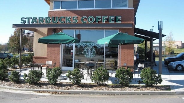 A Starbucks Coffee storefront with outdoor seating and a clear sky.