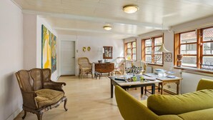 Ein geräumiges Wohnzimmer mit Holzboden, einem Esstisch und zwei Stühlen. Zwei Fenster mit Vorhängen, eine Tür und eine Wand mit zwei Bildern sind zu sehen.