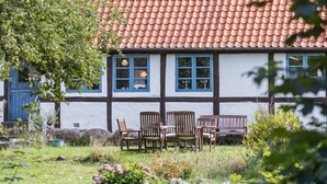Ein traditionelles Fachwerkhäuschen mit rotem Ziegeldach, ein Garten mit blühenden Pflanzen und eine Bank davor.