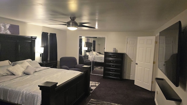 A bedroom with a bed, a chair, a dresser, a ceiling fan, and a TV on the wall.