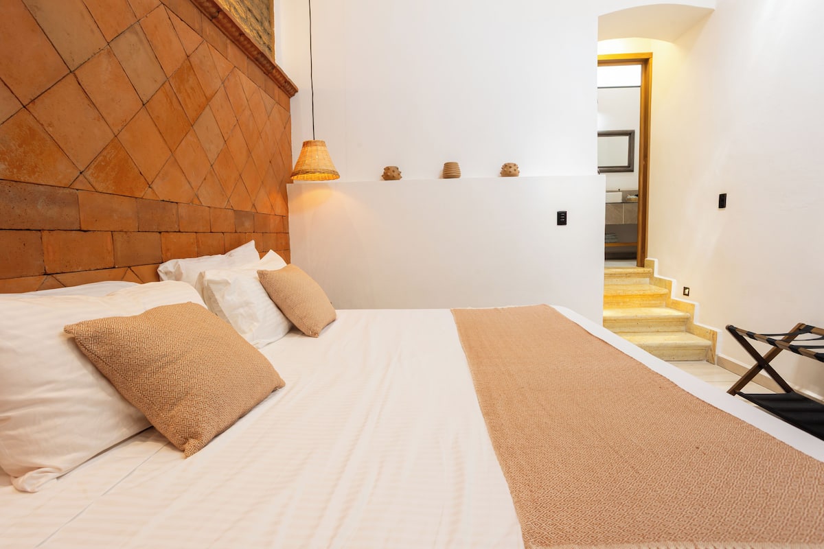 Quarto com cama, almofadas, luminária de cabeceira, cabeceira de madeira, prateleira pequena com objetos decorativos e escada que leva a um banheiro.