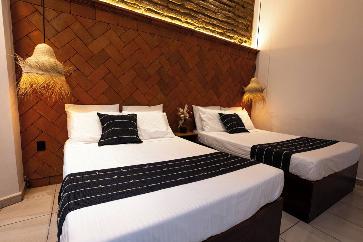 Quarto de hotel com duas camas, painel de cabeceira com padrão de tijolos e duas luminárias fixadas na parede.