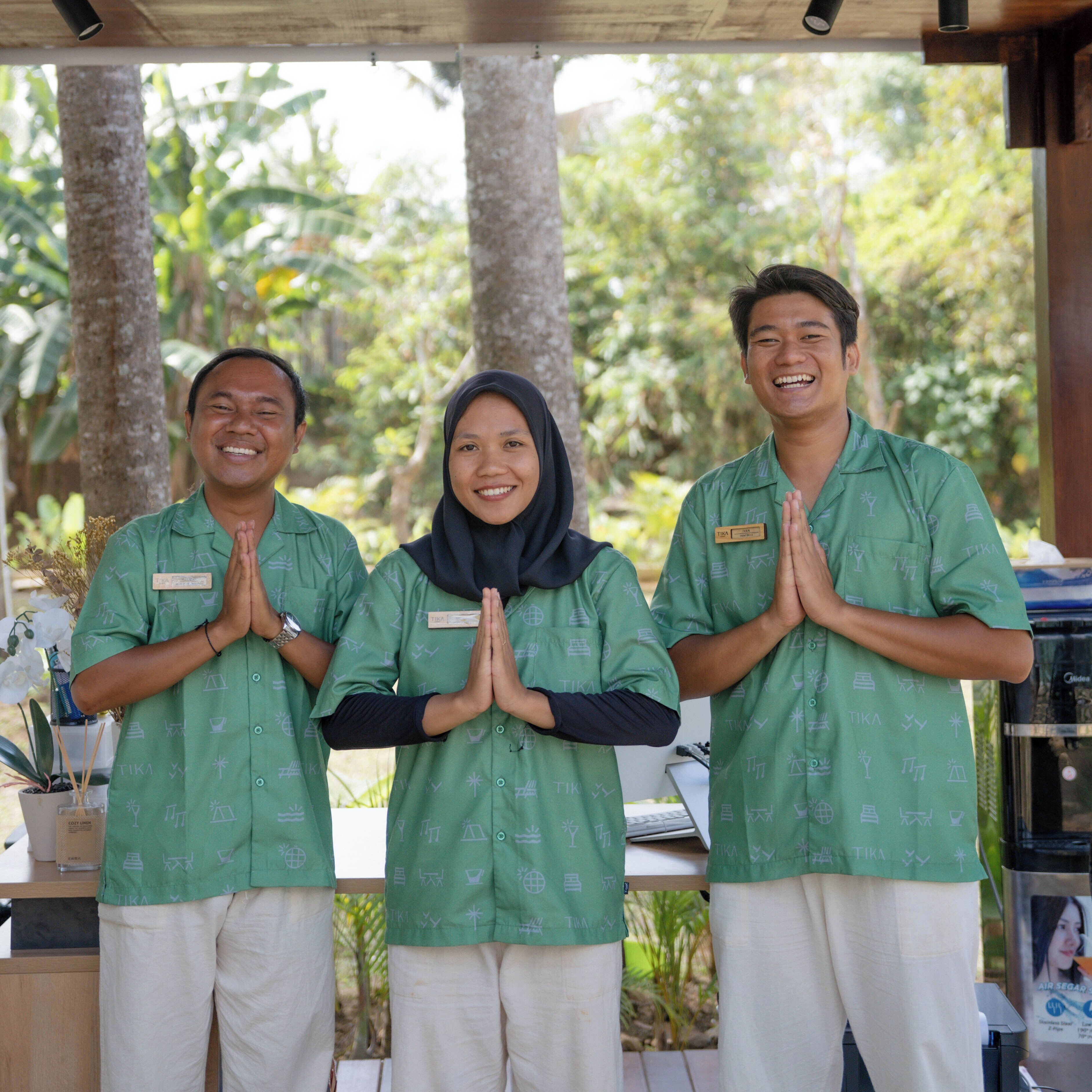 Three individuals in green uniforms standing in a line with their hands in a prayer position, outdoors with trees in the background.