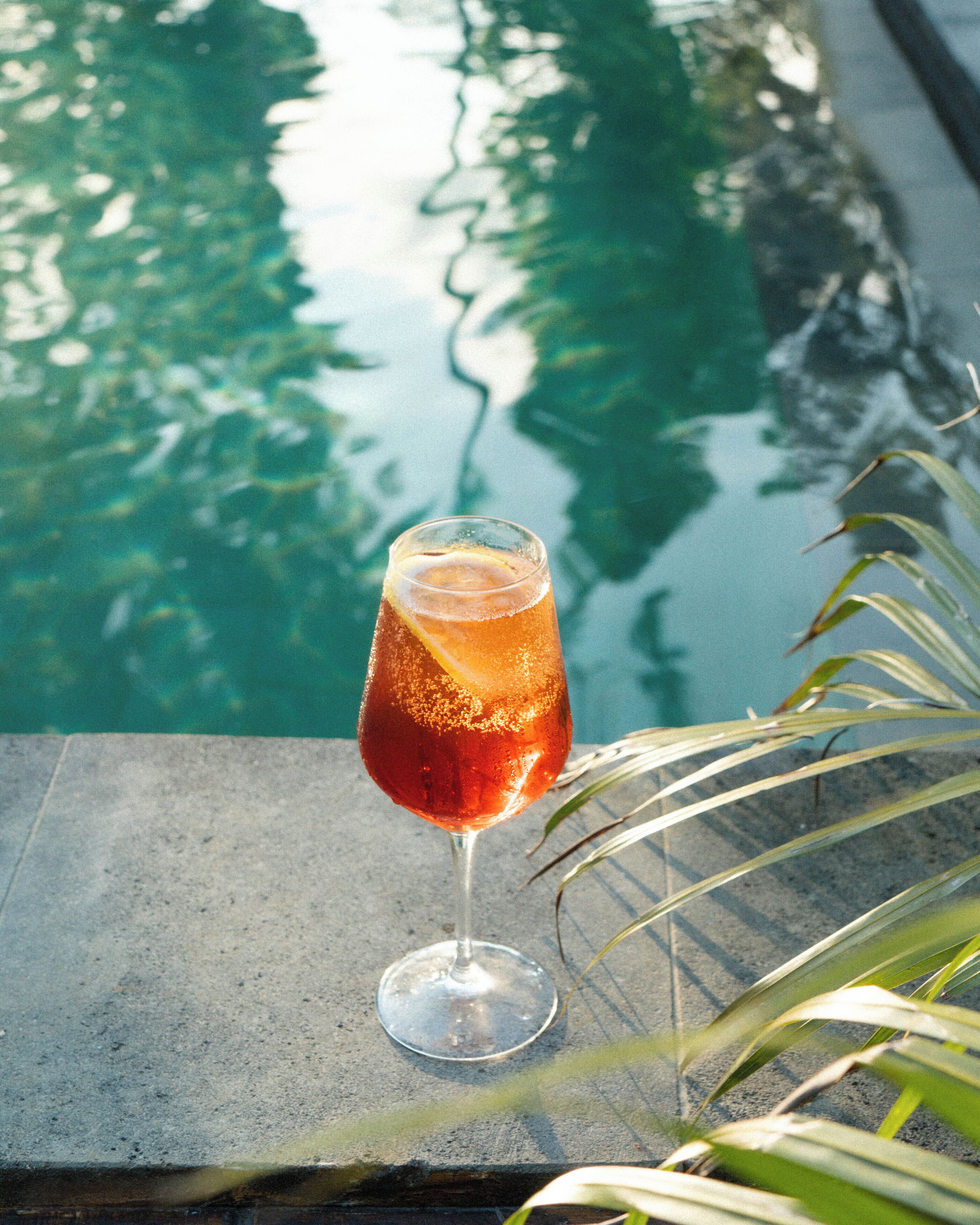 Un vaso de bebida de color ámbar con un gajo de limón sobre una superficie de piedra junto a una piscina.