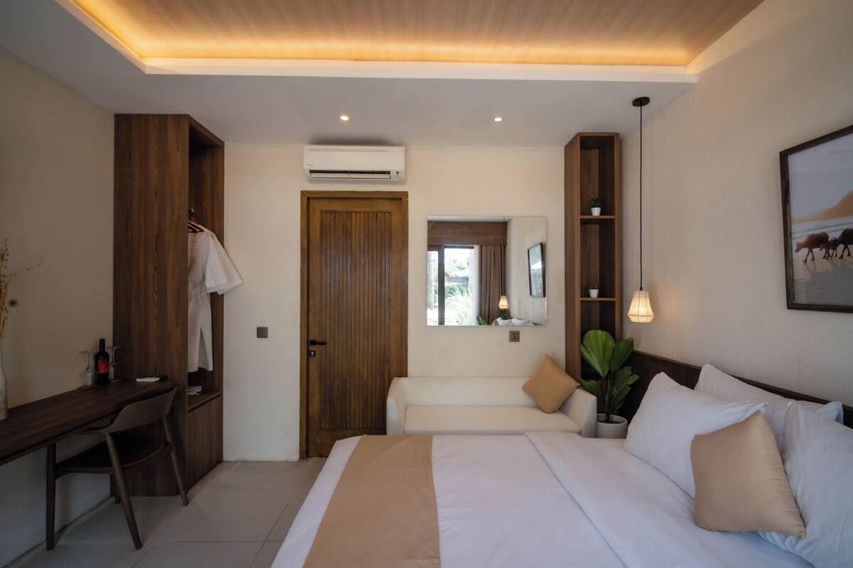 A hotel room with a bed, a desk, a chair, a wardrobe, a mirror, a plant, and a painting on the wall.