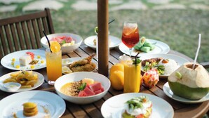 A dining table with various dishes and drinks, surrounded by chairs, set outdoors with a pool in the background. - Tika Lombok (Kuta)