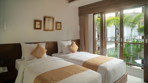 A hotel room with two beds, a wooden headboard, a telephone, and a view of greenery through large windows. - Tika Lombok (Kuta)