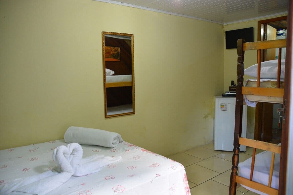 A single bed with a floral bedspread and a rolled towel, a small white refrigerator, and a wooden bunk bed in a room with plain walls and a tiled floor.