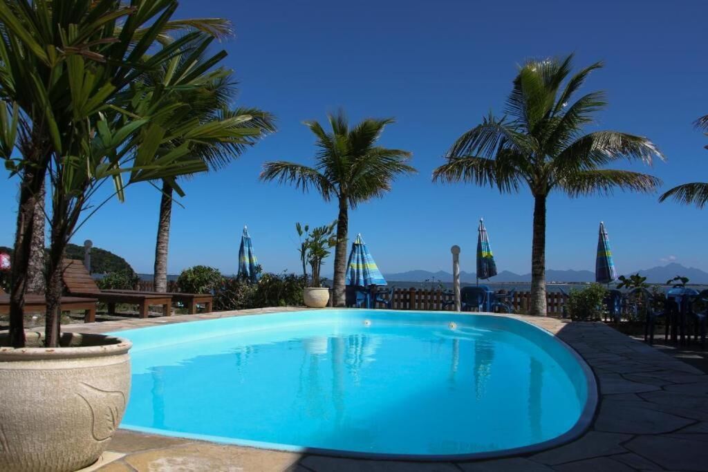 A clear blue swimming pool surrounded by palm trees and beach umbrellas, with a view of the ocean and mountains in the background.