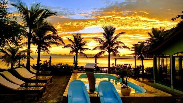 Zona de piscina con sillas de descanso y palmeras, con vista a la puesta de sol sobre el océano.