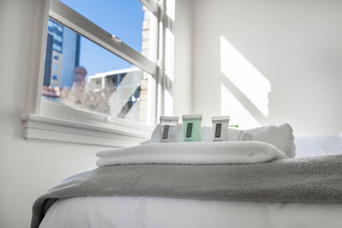 A neatly made bed with a grey blanket and a white towel, a window with a view of buildings, and three toiletries on the bed.