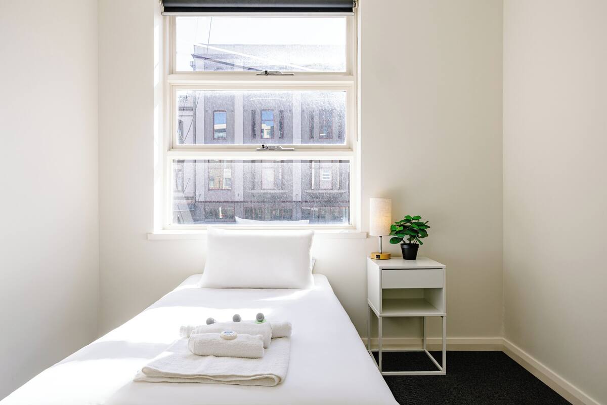 A hotel room with a bed, a nightstand with a lamp and a plant, and a window with a view of buildings.