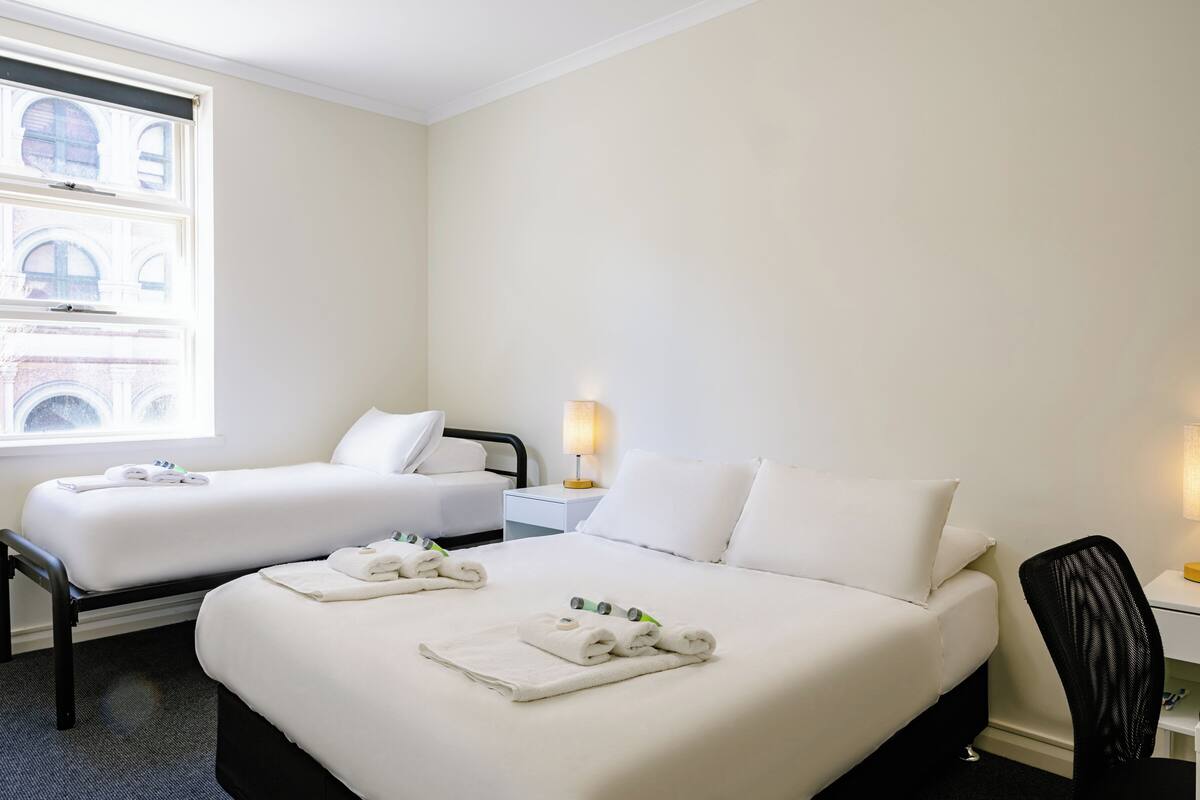 A hotel room with two beds, a desk, a chair, and a window with a view of buildings.