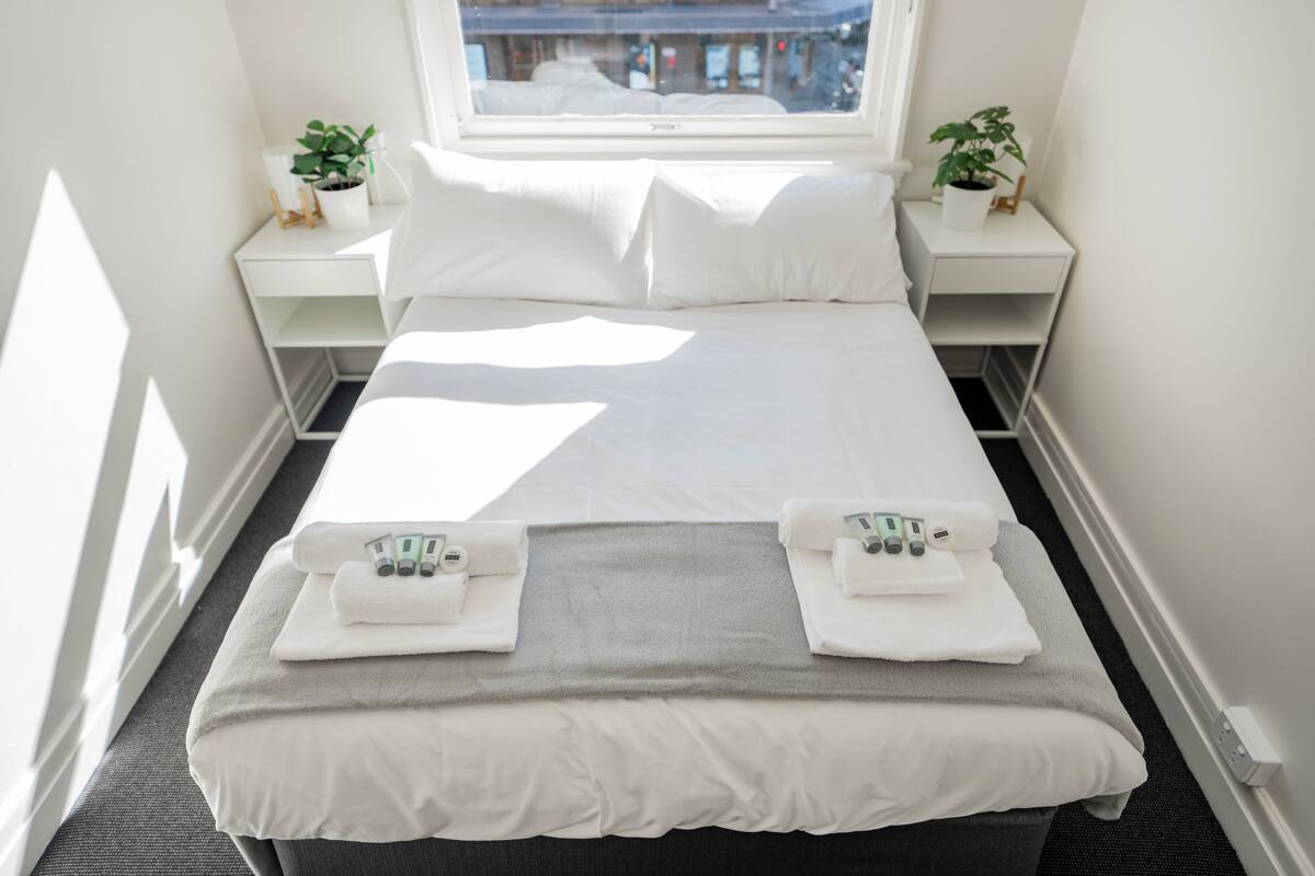A neatly made bed with white linens and towels, a grey blanket, and a window with a view of buildings.