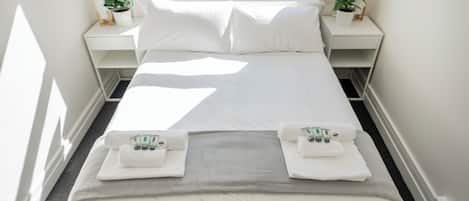 Cama bem arrumada com lençóis e toalhas brancos, um cobertor cinza e uma janela com vista para prédios.
