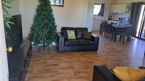 Un salon doté d’un plancher en bois, d’un canapé en cuir noir avec des coussins, d’un petit sapin de Noël et d’un coin repas avec une table et des chaises.