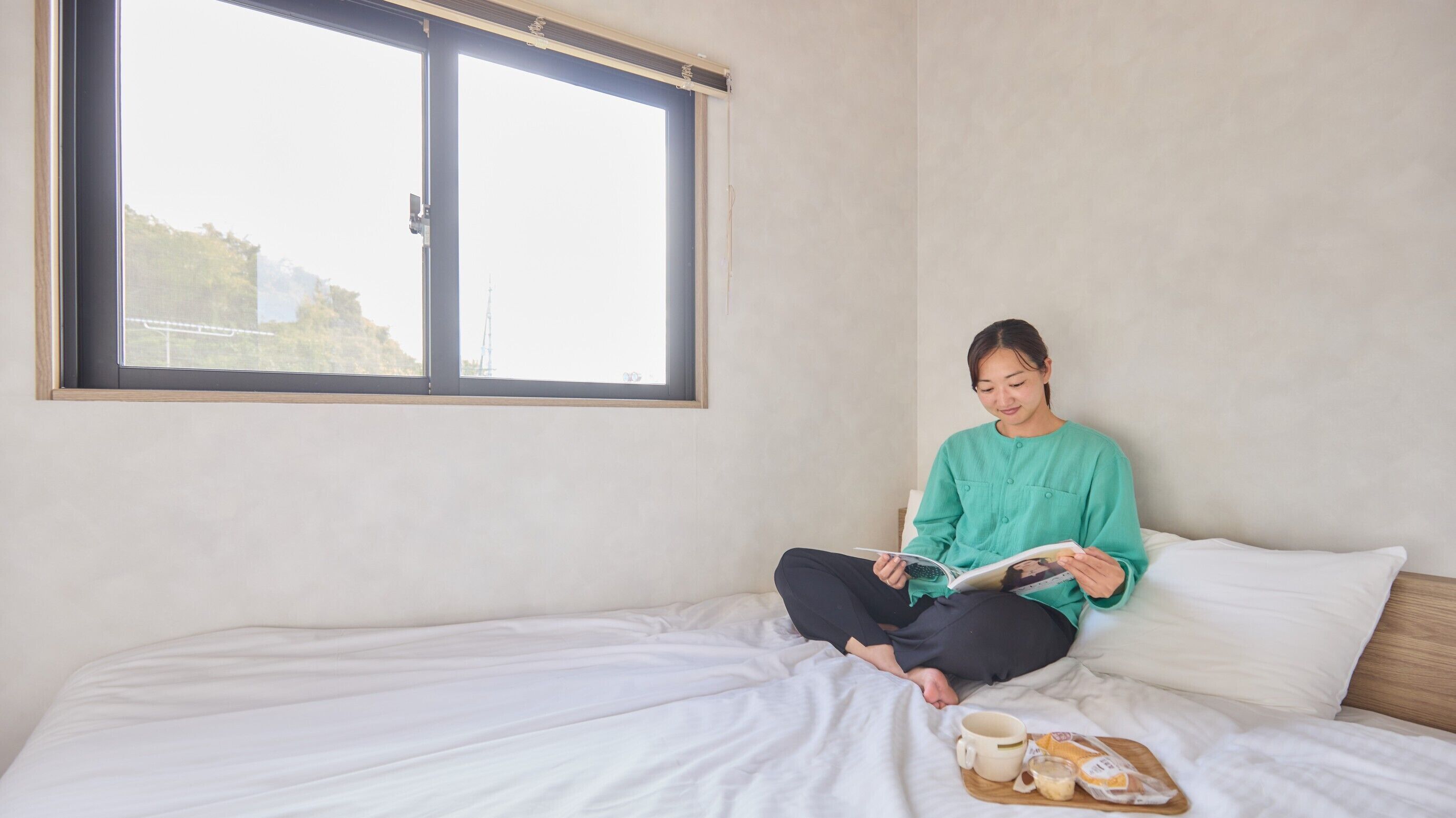 Una persona sentada en una cama, con una ventana cerca, leyendo un libro y una bandeja con comida y una taza.
