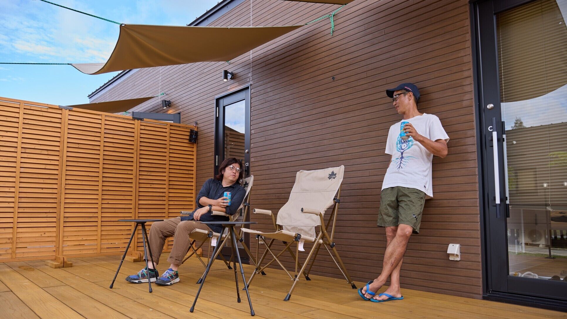 Una terraza de madera con dos sillas plegables, una mesa y una persona de pie junto a un ventanal corredizo.