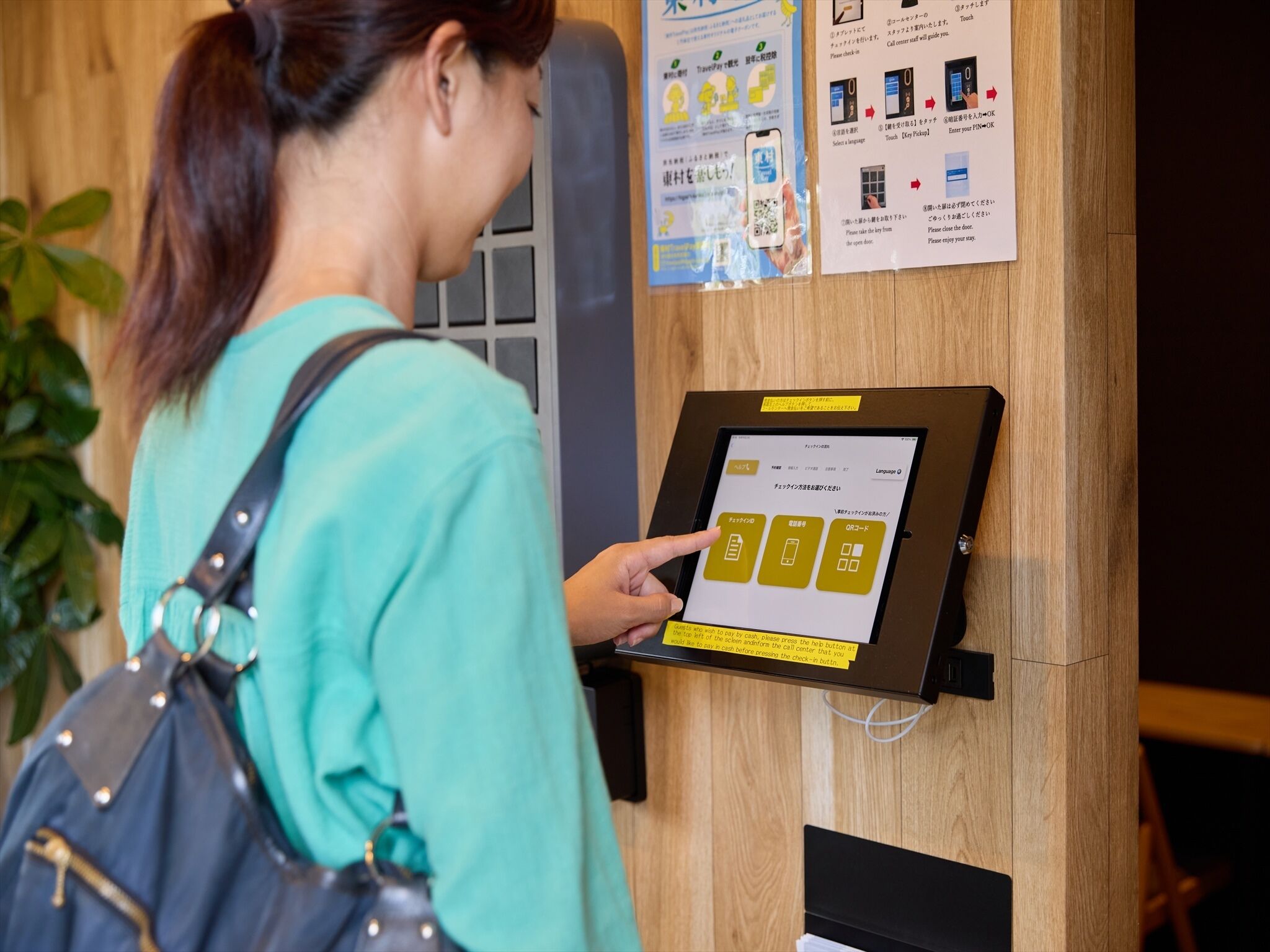 A woman interacting with a touchscreen kiosk, possibly for check-in or information, in a room with wooden paneling and a plant in the background.