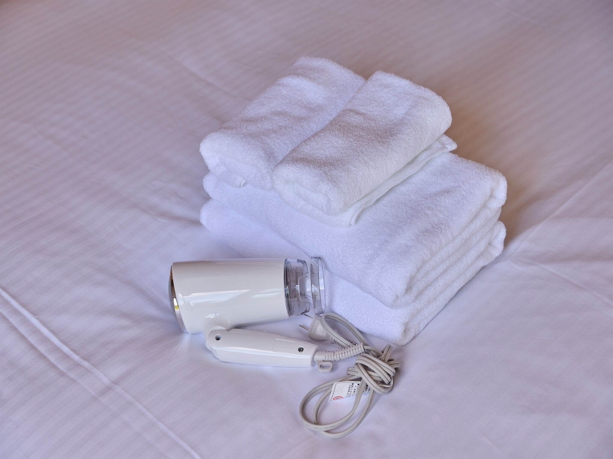 A neatly folded white towel and a portable hair dryer on a bed.