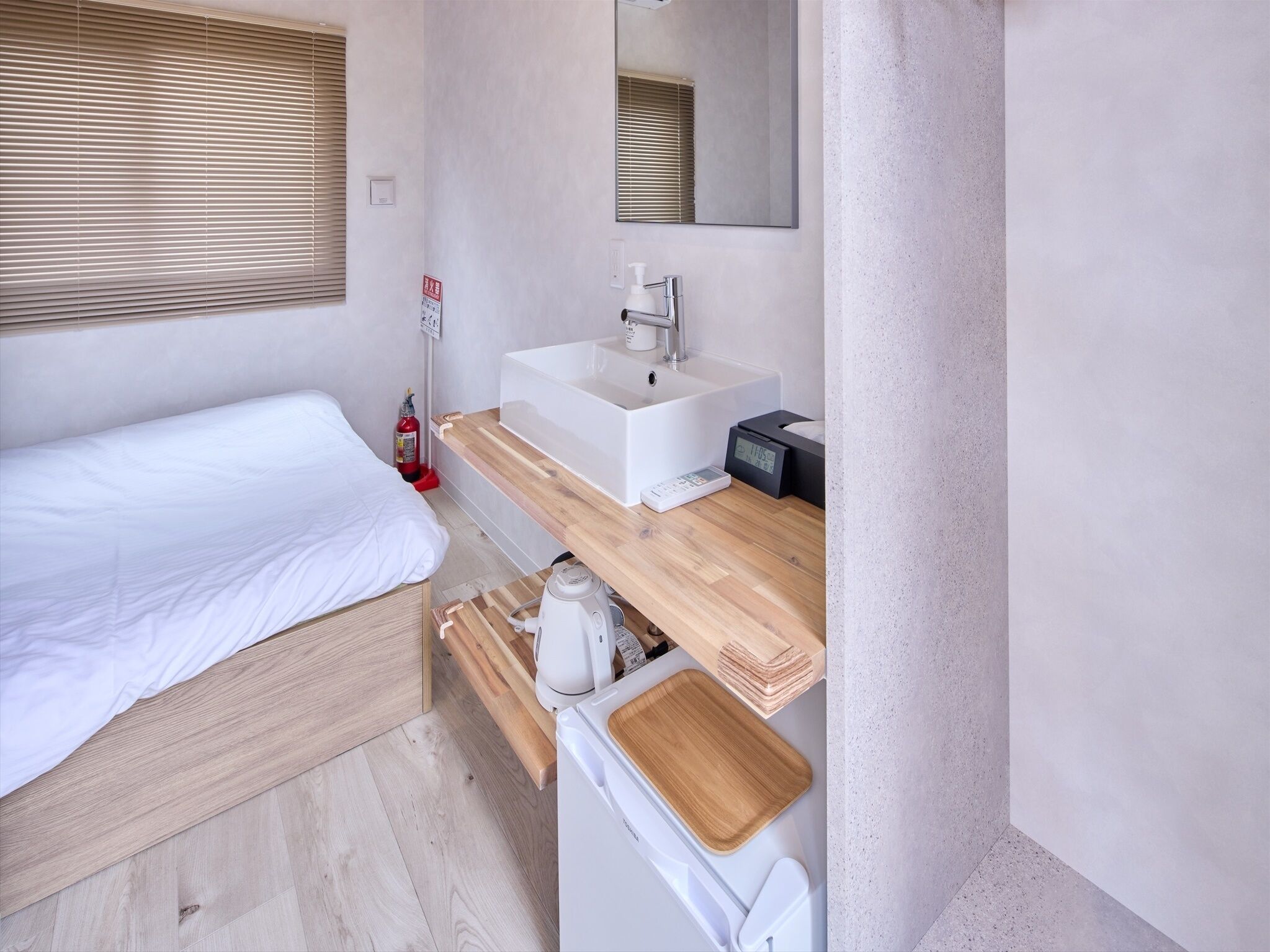 A modern bedroom with a bed, a white sink, a mirror, a wooden shelf, and a small kitchen area with a kettle and a toaster.