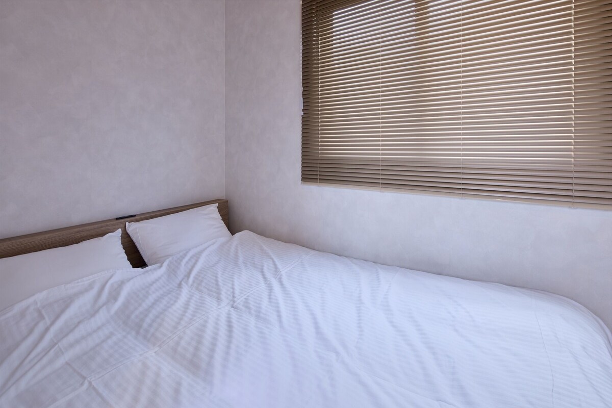 Un lit bien fait, avec du linge de lit blanc, dans une pièce aux murs unis et dotée d’une fenêtre avec des stores horizontaux.