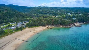 An aerial view of a coastal area with a beach, clear turquoise waters, and a forested hillside in the background. - Yambaru Experience Hotel Nunganiku (Higashi)
