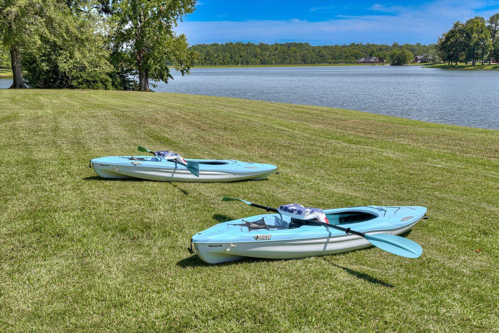 Two kayaks on a grassy area by a lake with trees and houses in the background.