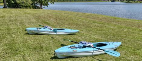 Two kayaks on a grassy area by a lake with trees and houses in the background.