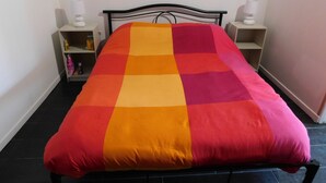 A bed with a checkered bedspread in red, orange, and yellow. There are two bedside tables with lamps and a shelf with items on the left side. - Short-term furnished studio rental (n° 3) (Le Trait)