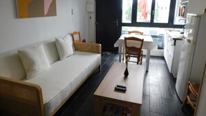 A living room with a sofa, a coffee table, and a dining area with a table and chairs. There is a painting on the wall and a window with curtains. - Short-term furnished studio rental (n° 3) (Le Trait)