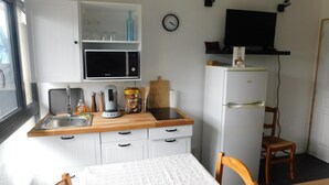 A compact kitchen with a microwave, coffee maker, and a refrigerator. There is a wooden table and chairs, a clock, and a window with blinds. - Short-term furnished studio rental (n° 3) (Le Trait)