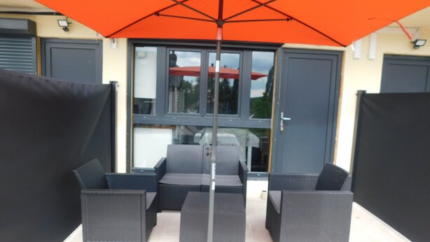 An outdoor seating area with a large orange umbrella, black wicker furniture, and a glass door entrance. - Short-term furnished studio rental (n° 3) (Le Trait)