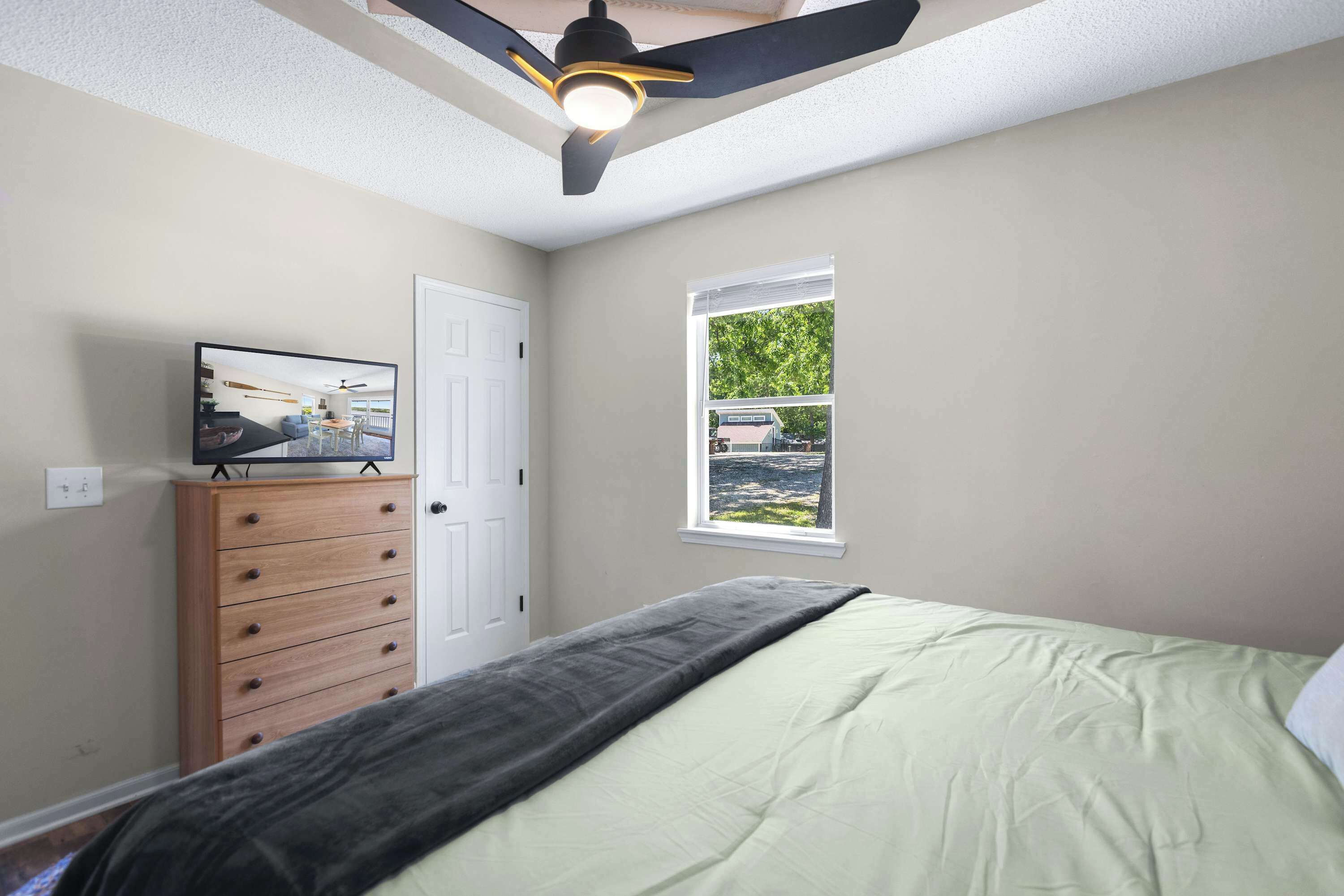 Une chambre à coucher avec un lit, une commode, un ventilateur de plafond, une fenêtre et un téléviseur posé sur la commode.