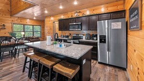 A modern kitchen with wooden walls, a granite countertop, stainless steel appliances, and a bar area with stools. - Modern Cabin w/ Pool, Hot Tub, Games & More! (Sevierville)