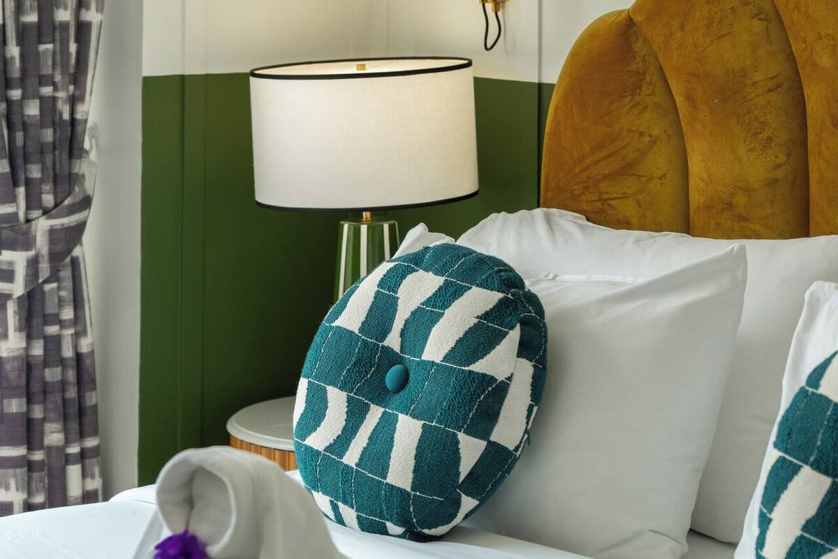 Une chambre d’hôtel avec un lit, une tête de lit verte, une lampe fixée au mur, une illustration botanique encadrée et un coussin à motifs.