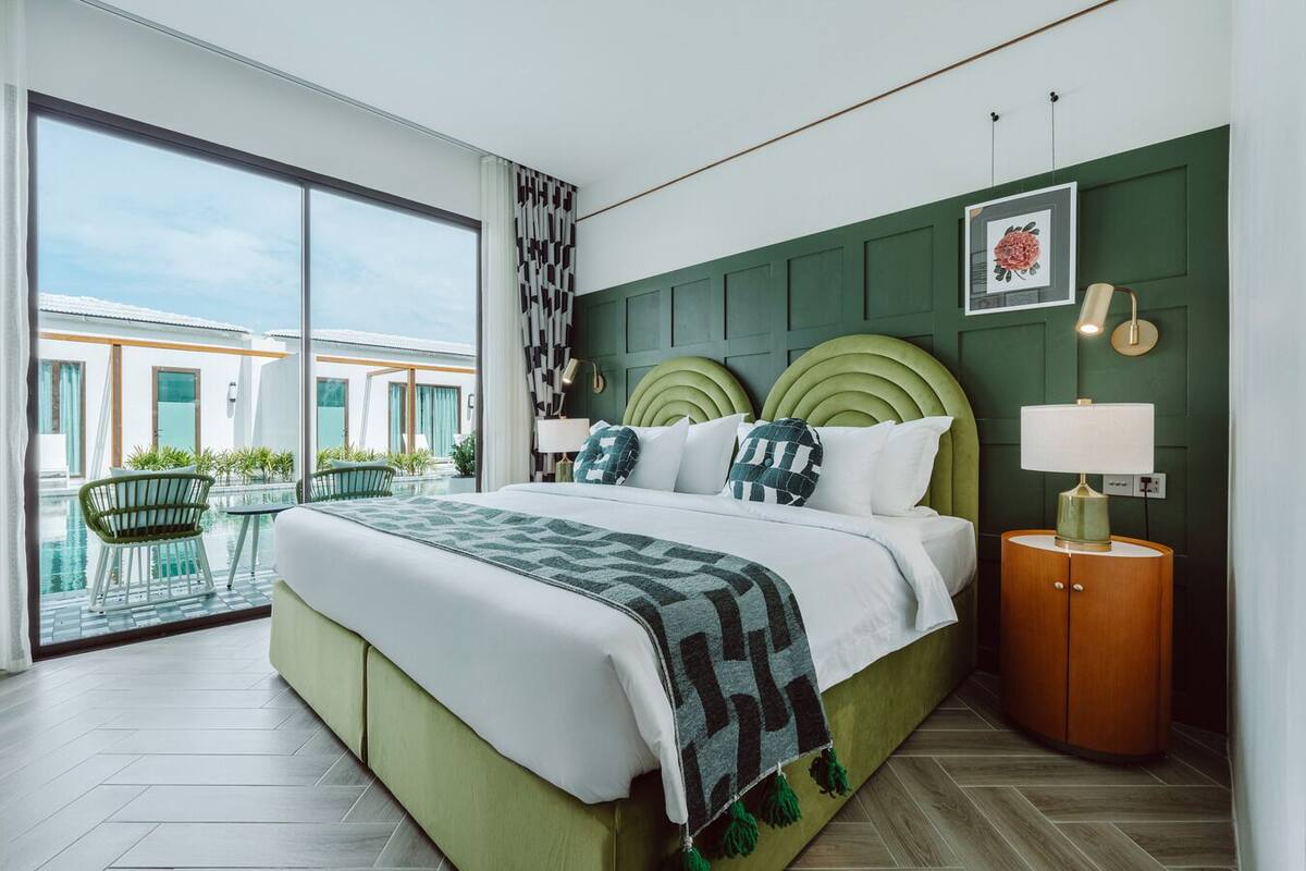 Une chambre d’hôtel moderne avec un grand lit, une tête de lit verte, une table de chevet avec une lampe, et une vue sur une piscine et un espace de détente extérieur.