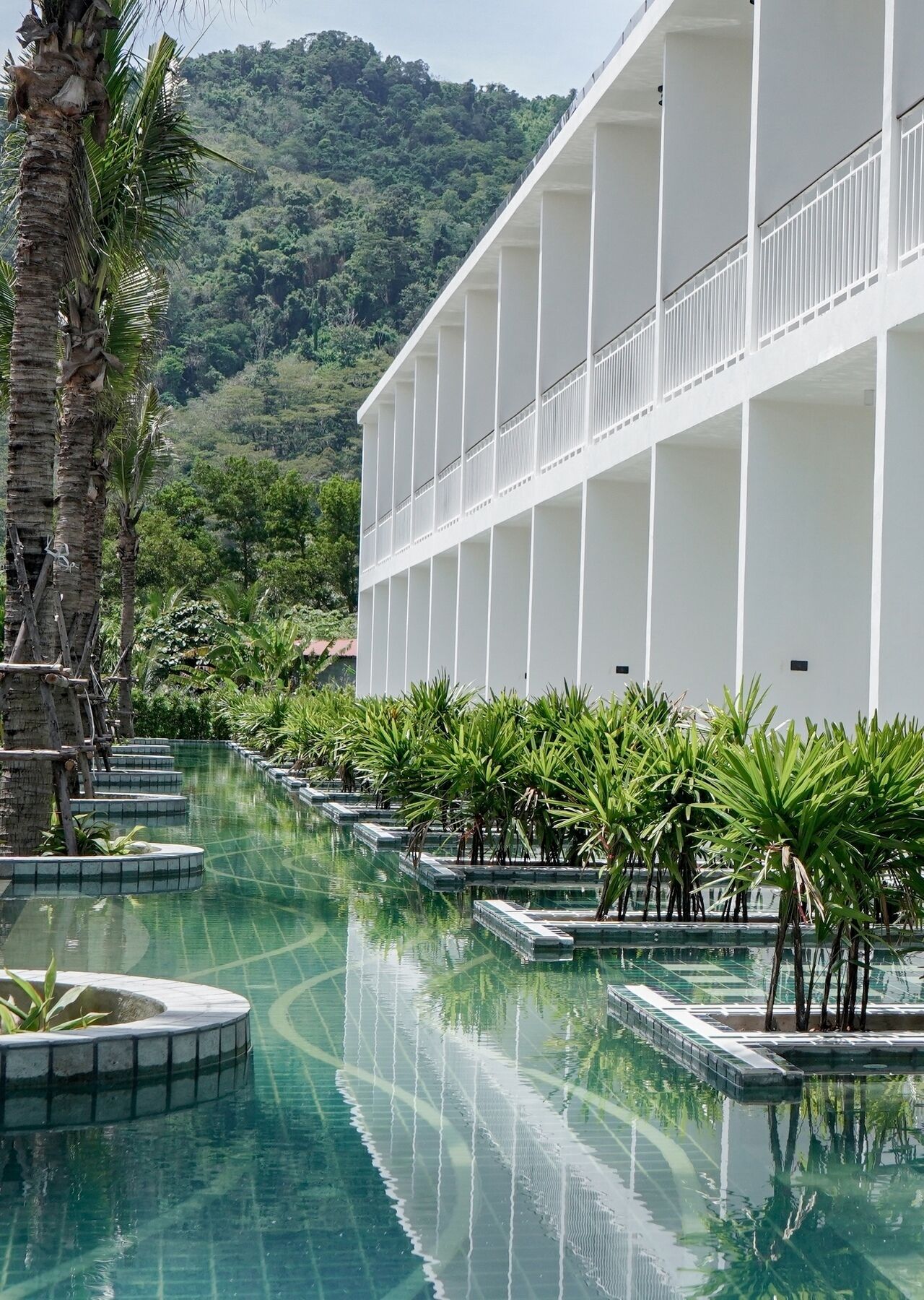 A pool area with a modern building, palm trees, and a mountainous backdrop.