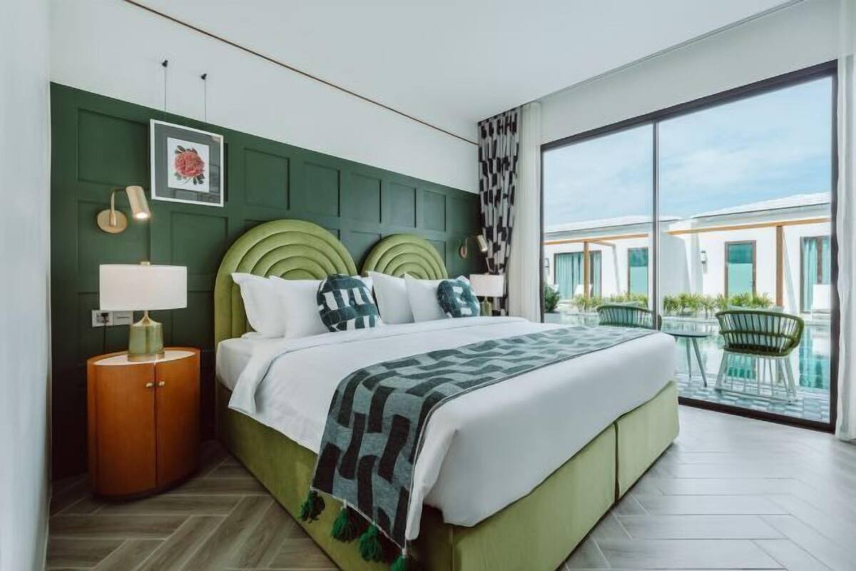 Une chambre d’hôtel moderne avec une tête de lit verte, un grand lit, une table de chevet avec une lampe et une vue sur l’espace extérieur.