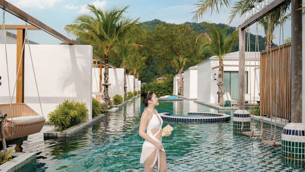Une femme se tient dans une piscine, avec vue sur un complexe hôtelier comprenant une structure en bois et des palmiers.