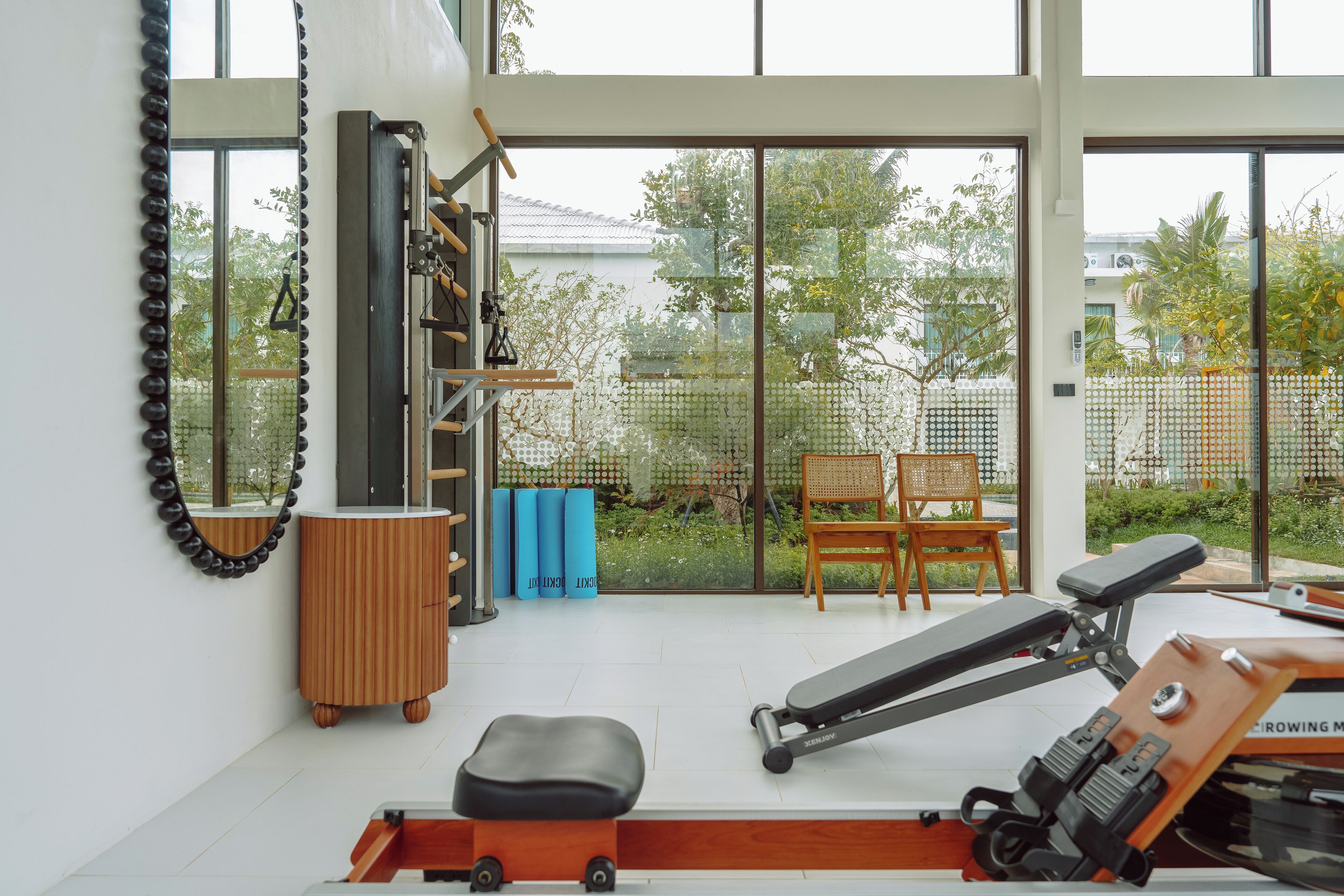 A modern gym with exercise equipment, a wooden bench, and a view of a garden through large windows.