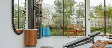 A modern gym with exercise equipment, a wooden bench, and a view of a garden through large windows.