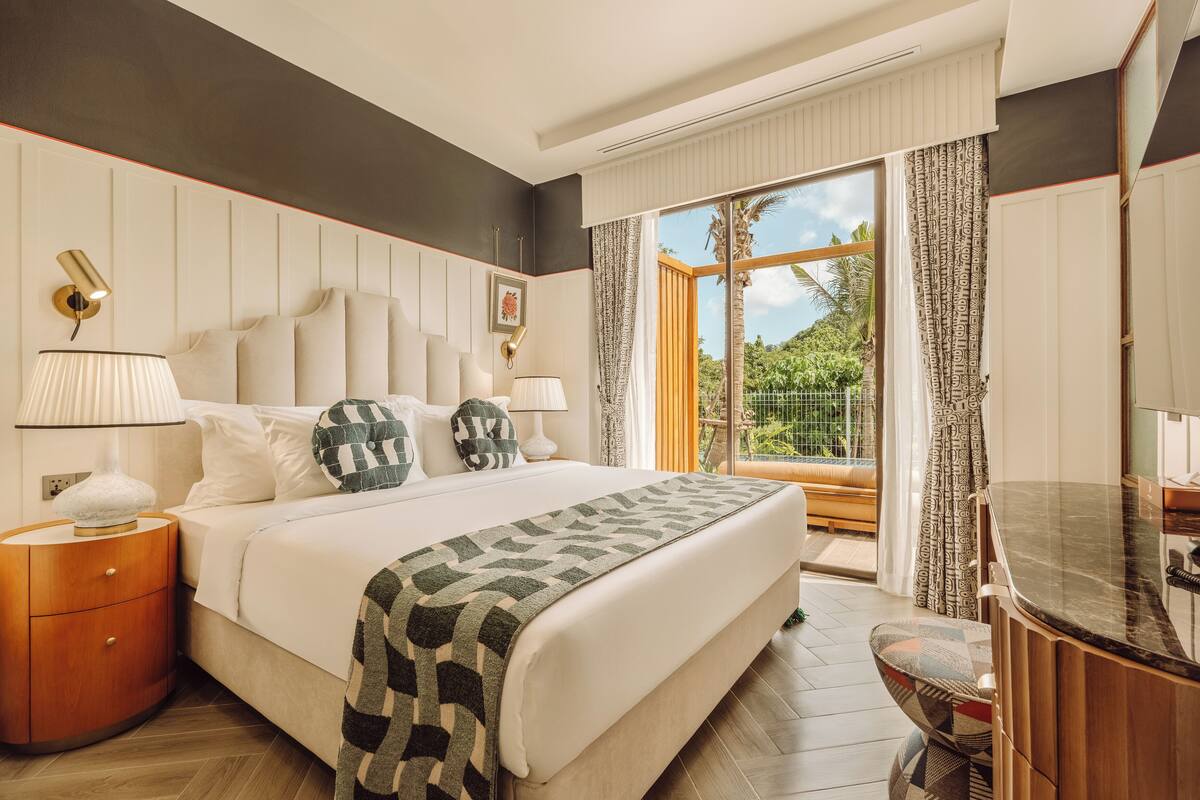 Une chambre d’hôtel moderne avec un grand lit, une table de chevet en bois, un plan de toilette en marbre dans la salle de bain et un balcon offrant une vue sur la verdure.