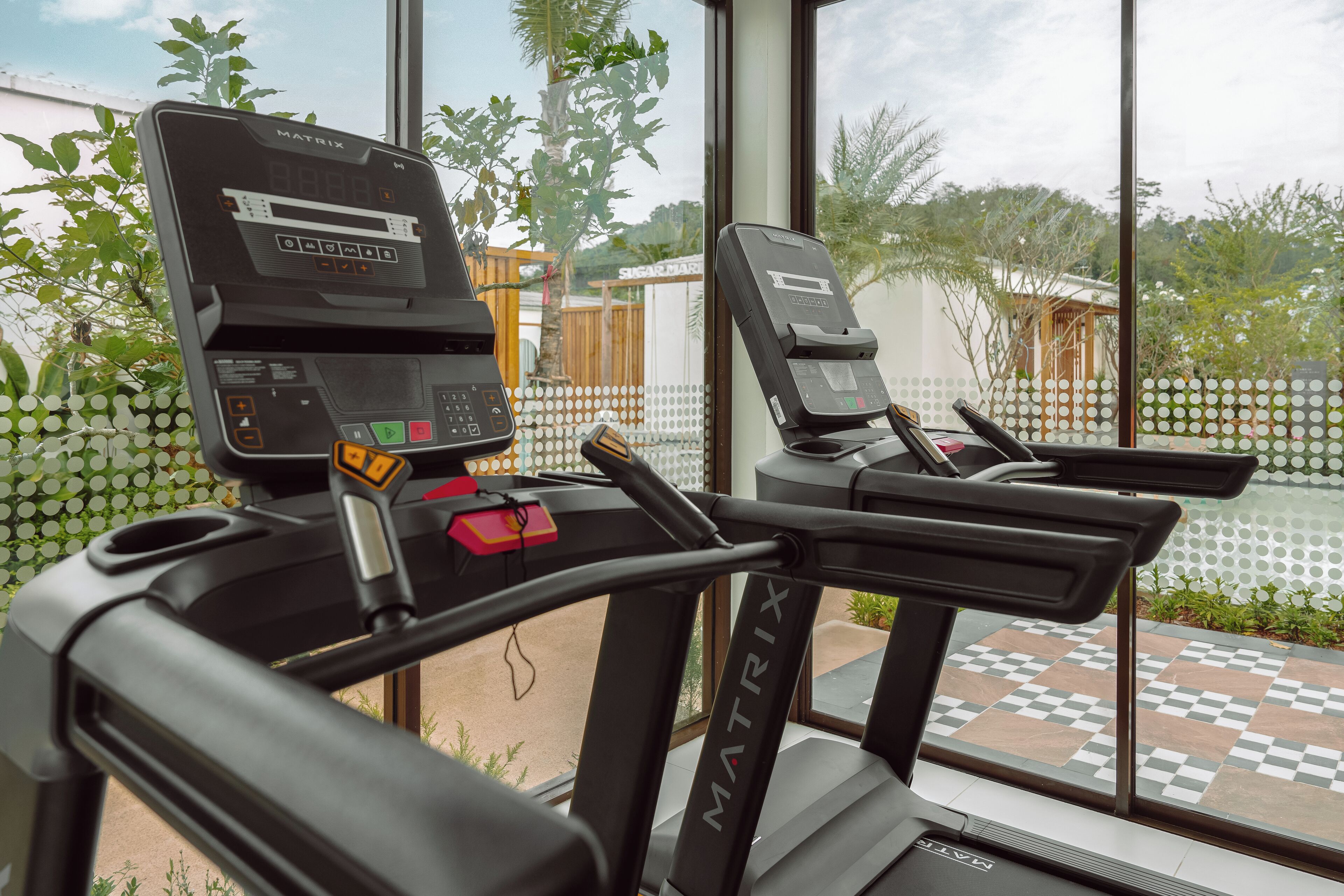 A treadmill with a digital display and control panel, positioned near a window with a view of a garden and outdoor area.