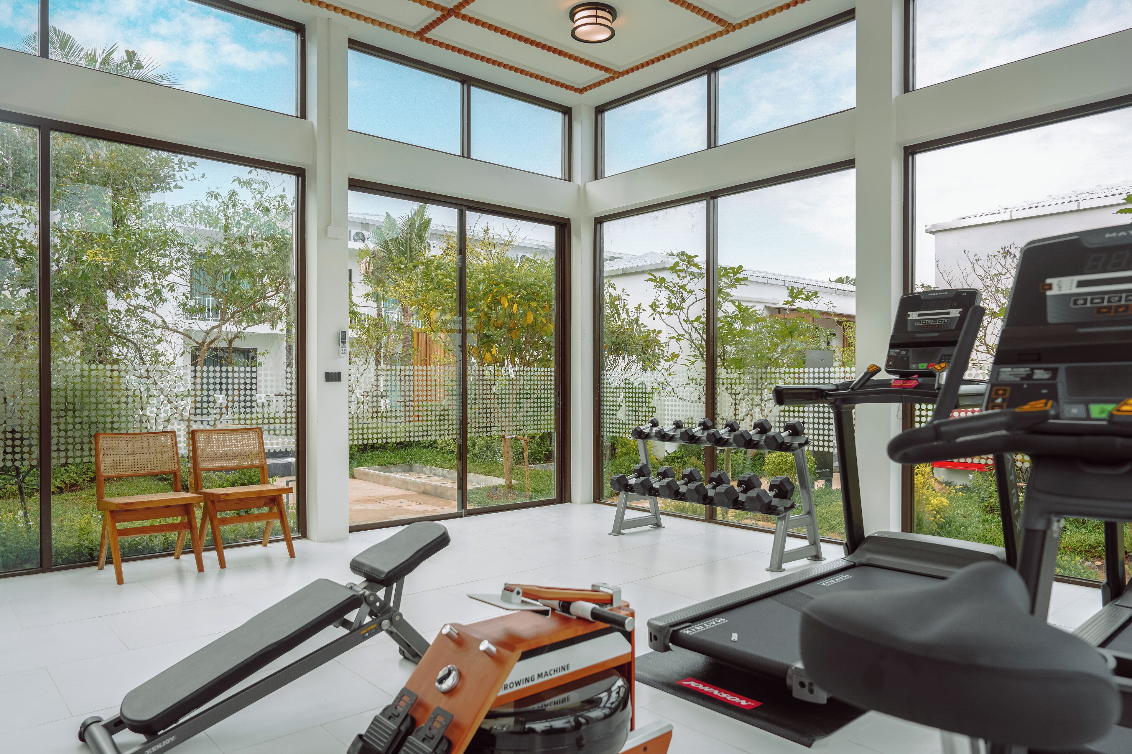 A spacious gym with large windows, a treadmill, a rowing machine, dumbbells, and two wooden chairs.